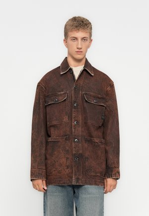 Jonge man in een vervaagde donkerbruine knoopjasje met grote klepzakken en lichtblauwe jeans tegen een plain witte achtergrond.