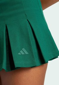 Zelená skladaná sukňa vyrobená z textúrovanej tkaniny, s malým vyrazeným logom Adidas v dolnom rohu.