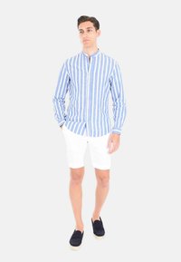 Camicia a maniche lunghe a strisce blu e bianche con colletto alla cinese, abbinata a pantaloni corti bianchi e scarpe slip-on blu scuro. Sfondo semplice.