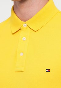 Gros plan d'un polo jaune avec trois boutons blancs et un petit logo rectangulaire rouge, blanc et bleu sur la poitrine.