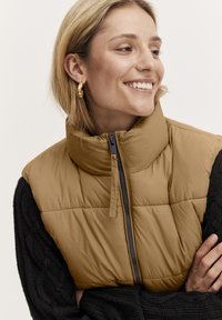 Khakifarbene puffy Weste mit hohem Kragen, Reißverschluss vorne und gesteppter Optik; kombiniert mit einem strukturierten schwarzen Kabelstrickpullover und goldenen Creolen.