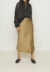 Haut en satin noir avec manches en dentelle et col à nouer, associé à une jupe midi en satin doré. Des chaussures plates noires complètent la tenue.