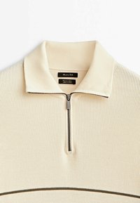 Pull beige clair à fermeture éclair avec texture côtelée, détail rayé foncé contrastant et col polo classique. Doté d'une fermeture éclair en métal argenté.