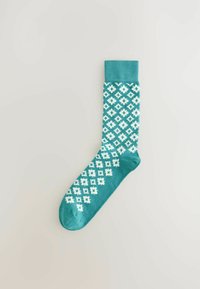 Un calcetín en color verde azulado con un patrón geométrico en blanco de cuadrados y rombos. Puño acanalado, textura suave y un diseño de punta reforzada.