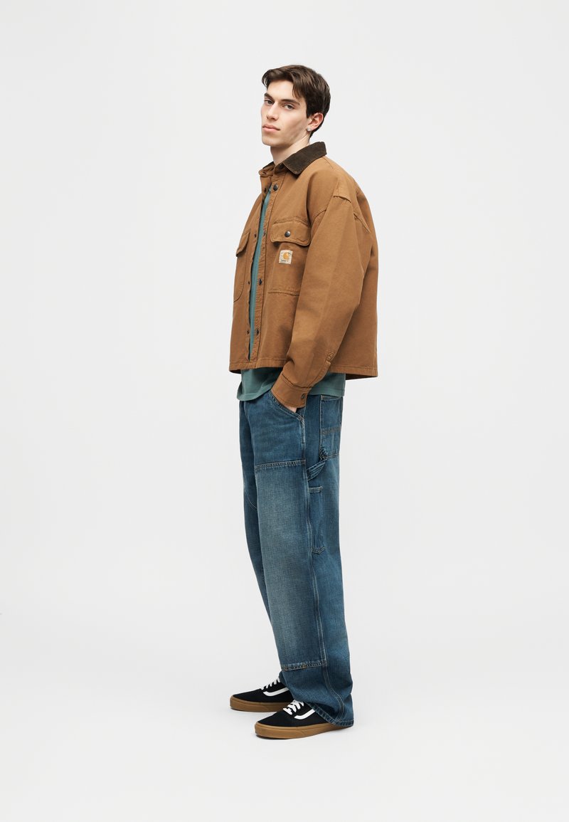 Junger Mann in brauner Jacke, türkisfarbenem Hemd, lockeren blauen Jeans und schwarzen Turnschuhen vor einem schlichten weißen Hintergrund.