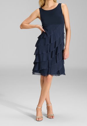 Femme portant une robe bleu marine sans manches avec des volants superposés et des talons ouverts argentés, debout avec une main sur la hanche sur un fond uni.