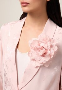 Lichtroze satijnen blazer met een bloemenkant broche. Gedetailleerd bloemenpatroon op de stof, zachte textuur en elegant ontwerp met sjawlcollar.