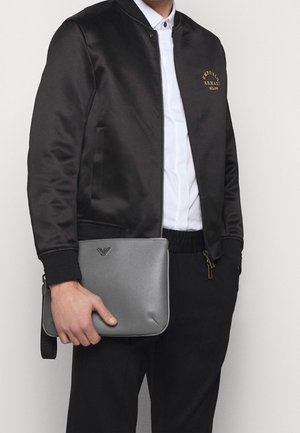 Satin-schwarze Bomberjacke mit goldenem Logo, kombiniert mit einer grauen strukturierten Dokumententasche mit Logo und seitlichem Griff.