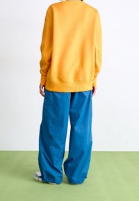 Gul sweatshirt med en avslappnad passform, i kombination med lösa, blå cargobyxor med sidofickor, mot en enfärgad bakgrund.