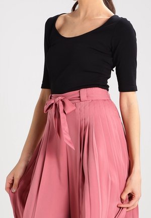 Femme portant un haut noir à manches mi-longues et une jupe plissée rose taille haute, ceinturée d'un nœud assorti.