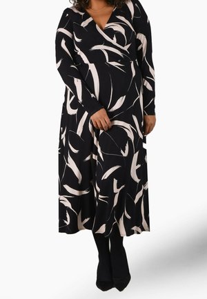 Kobieta ubrana w czarną sukienkę midi na zakładkę z białymi abstrakcyjnymi wzorami pędzla, długimi rękawami i czarnymi butami z noskiem w szpic.