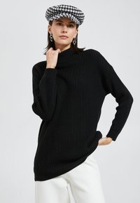 Schwarzer Strickpullover mit hohem Kragen, legerem Schnitt und gerippter Textur. Das Model trägt eine karierte Mütze und weiße Hosen.