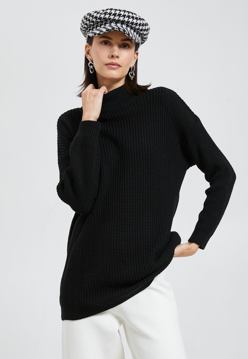 Schwarzer Strickpullover mit hohem Kragen, legerem Schnitt und gerippter Textur. Das Model trägt eine karierte Mütze und weiße Hosen.
