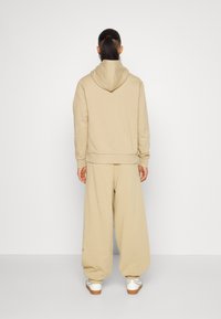Beige hoodie met een relaxte pasvorm, gecombineerd met bijpassende wijde sweatpants. Soepele textuur en geribbelde manchetten. Witte sneakers zichtbaar.