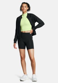 Giacca bomber leggera nera sopra un crop top aderente verde lime e pantaloni corti neri a vita alta, abbinati a scarpe da ginnastica bianche.