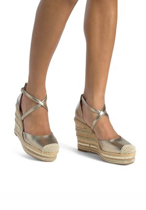 Metalen gouden wedge sandalen hebben kruisbanden en een geweven juten platform. De neuskap is crèmekleurig, wat contrast toevoegt aan het ontwerp.