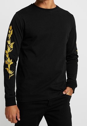 Homme portant une chemise noire à manches longues avec une broderie florale jaune le long de la manche droite, main dans la poche.