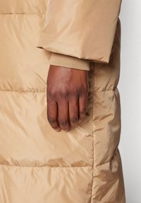Main à la peau foncée reposant contre la manche d'une veste matelassée beige avec détail de bord-côte, gros plan sur la texture du tissu et les coutures.