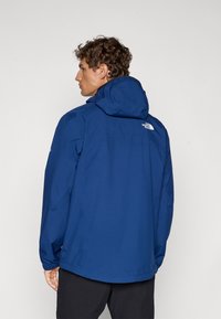 Σκούρο μπλε αδιάβροχο μπουφάν με κουκούλα, ελαστικούς καρπούς και μια διακριτική λογότυπο στην πλάτη. Λείο υλικό κατάλληλο για υπαίθριες δραστηριότητες.