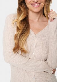 Vaaleanbeige neuletakki, jossa on V-pääntie, nappikiinnitys, ribbitekstuuri hienovaraisilla kuvioilla ja pitkät hihat.