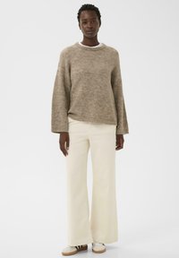 Femme portant un pull en tricot beige ample, un pantalon large crème et des baskets blanches avec des rayures noires sur fond blanc.