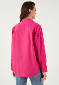 Long-sleeve shirt in vibrant pink, made from cotton fabric. Features a classic collar and button cuffs, with a relaxed silhouette and rounded hem.