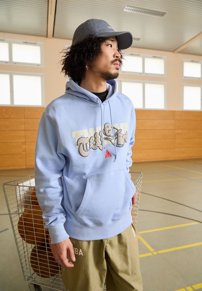 Sudadera azul claro con un gran gráfico en crema y gris, bolsillo tipo canguro, combinada con pantalones caqui que presentan texto y un logo.