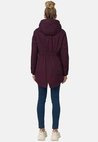 Long burgundy jacket with a hood, drawstring waist, and snap closures. Worn over blue jeans and light-colored sneakers.
