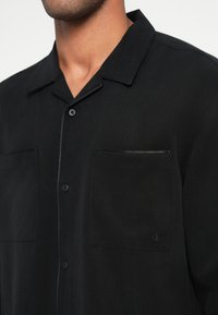 Musta lyhythihainen paita teksturoidusta kankaasta, jossa on napitettava kaulus, kaksi taskua ja hienovarainen logo vasemmassa rinnassa.