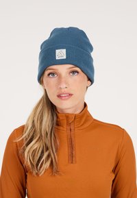 Blauwe gebreide beanie met een omgevouwen manchet en een vierkante logo patch. Gemaakt met een bruine hoog gesloten top met een rits, tegen een lichte achtergrond.