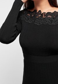 Pull noir côtelé avec un collier en dentelle détaillé. Les manches longues présentent un subtil détail bouffant. Tissu en mailles doux, design ajusté.