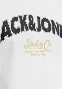 Camiseta de algodón blanca con texto en negrita negro "BLACK&JONES" y texto en dorado "Studio Co. Creado en Copenhague Desde mil novecientos noventa."