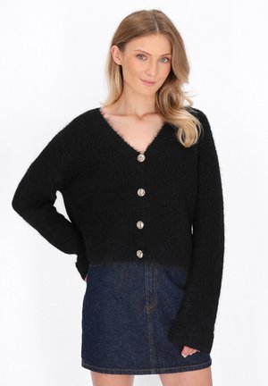 Schwarze kurze Strickjacke aus strukturiertem Strickstoff, mit einem V-Ausschnitt und sechs metallischen Knöpfen auf der Vorderseite. Kombiniert mit einem Jeansrock.