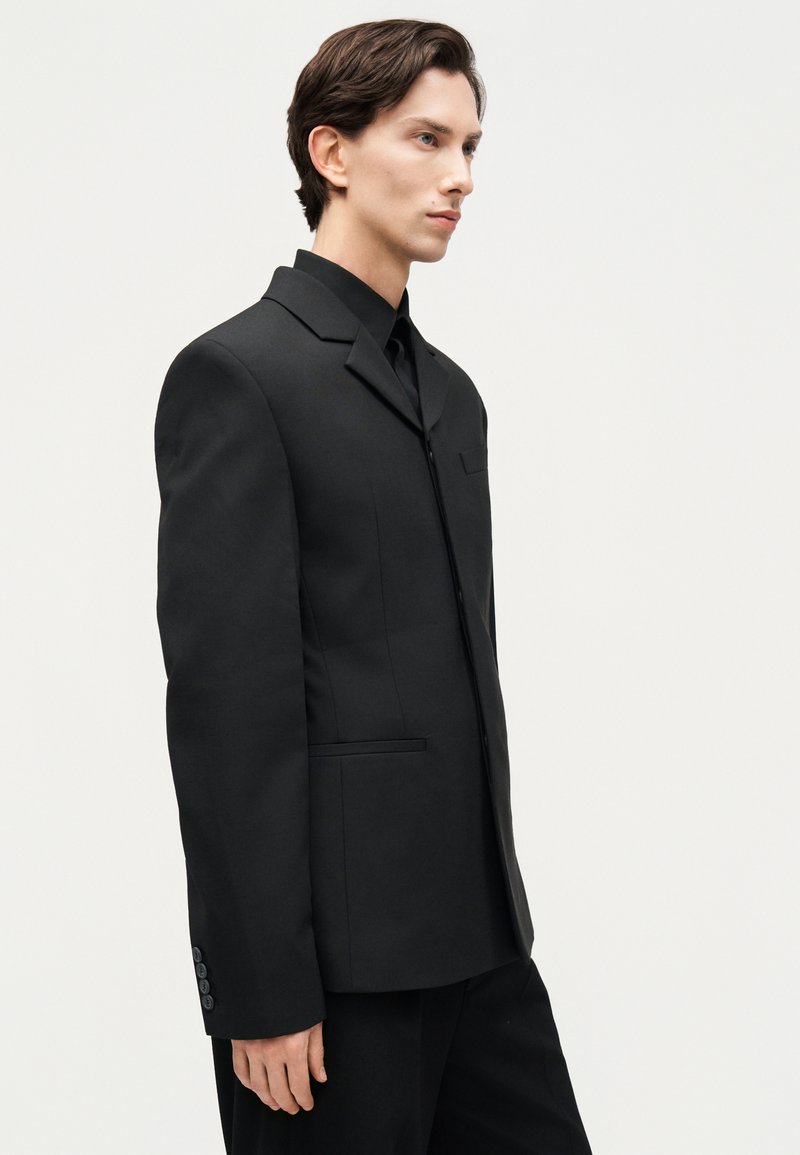 Mand iført sort skræddersyet blazer og matchende bukser, stående sidelæns mod en ensartet lys baggrund med neutralt ansigtsudtryk.