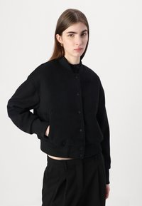 Giacca bomber nera realizzata in tessuto morbido, con chiusura a bottoni, polsini e orlo a costine. Il design ha una vestibilità comoda e dettagli minimi.