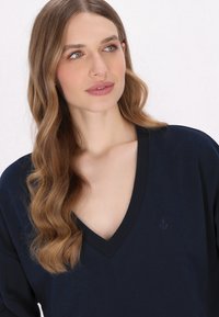 Námořnická modrá v-výstřihová halenka s jemnou texturou a žebrovaným výstřihem. Obsahuje vyšívaný detail kotvy na levé straně.