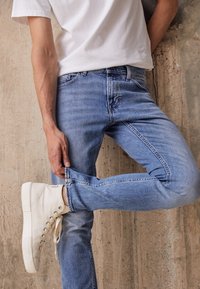 Una persona que lleva una camiseta blanca, jeans azules y zapatillas blancas posa frente a una pared gris texturizada, ajustándose el bajo de sus jeans.