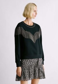 Felpa nera con frange e perline metalliche su un design a forma di V sul petto, indossata sopra una gonna plissettata con motivo.