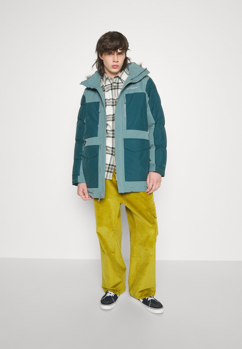 Veste matelassée turquoise et bleu clair avec de grandes poches, portée sur une chemise à carreaux et un pantalon en corduroy moutarde, accompagnée de baskets noires.