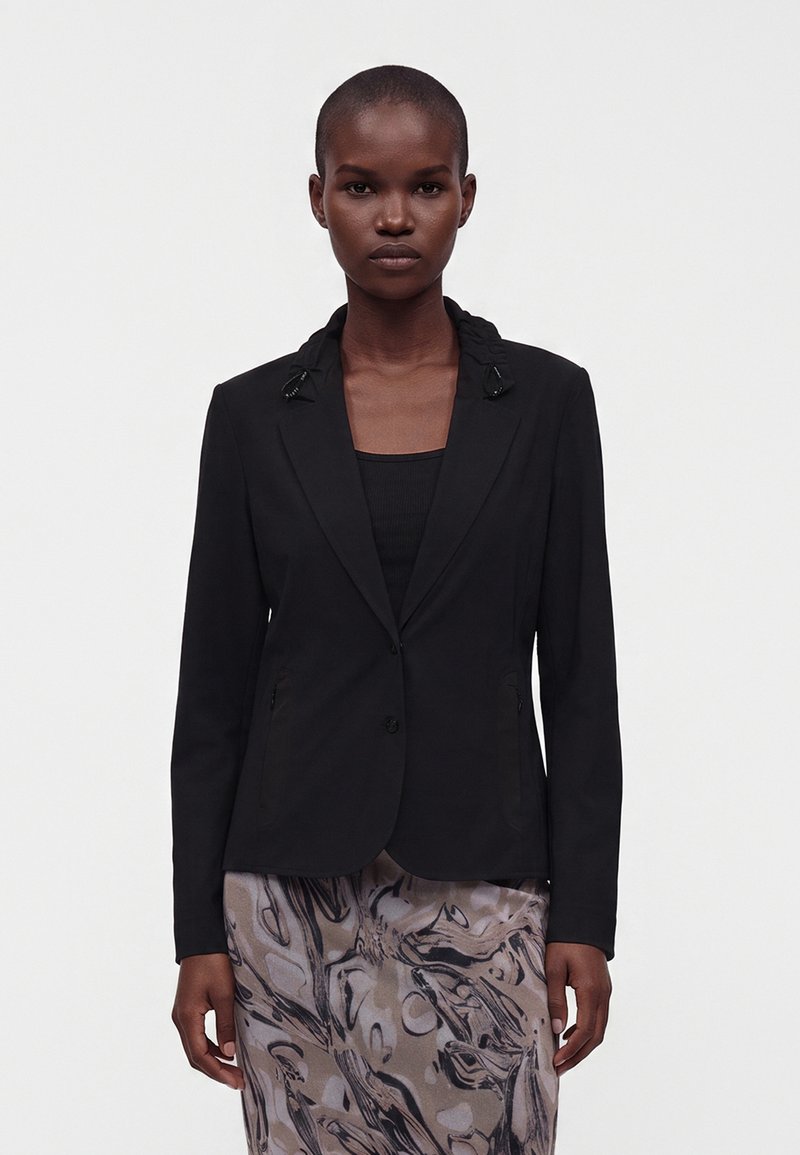 Femme portant un blazer noir sur un top noir et une jupe avec des motifs abstraits gris et noirs, se tenant devant un fond blanc uni.