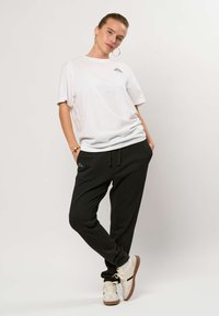 Kvinne står med hendene i lommer iført hvit t-skjorte med logo, svarte joggebukser, hvite joggesko og store hoop-ører mot ensfarget bakgrunn.