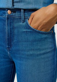 Vaqueros de mezclilla azul con textura descolorida, talle alto y detalle de bolsillo delantero. Cierre de botón de metal y costuras visibles.