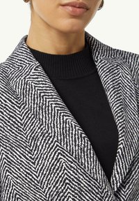 Schwarz-weißer Blazer mit Zickzack-Muster und fallendem Kragen, getragen über einem schwarzen, figurbetonten Pullover. Strukturierter Stoff mit einer klaren Silhouette.