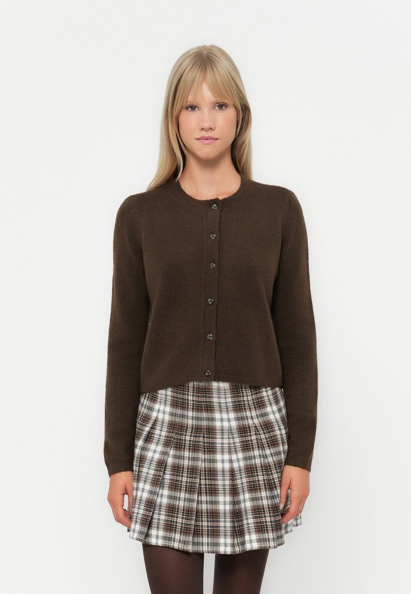 Καφέ κοντό σακάκι με στρογγυλή λαιμόκοψη και επτά πράσινα κουμπιά, φορεμένο πάνω από φούστα με πτυχές σε καρό σχέδιο που περιλαμβάνει λευκά, καφέ και μαύρα μοτίβα.