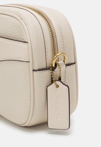 Krémová taška z texturované kůže s zlatými doplňky, která má horní zip a odnímatelnou visačku na korálkovém řetízku.