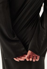 Zwarte satijnen jurk met lange, uitlopende mouwen. De stof is glad en glanzend, met een gedrapeerd ontwerp. De handen zijn achter de rug gekruist.