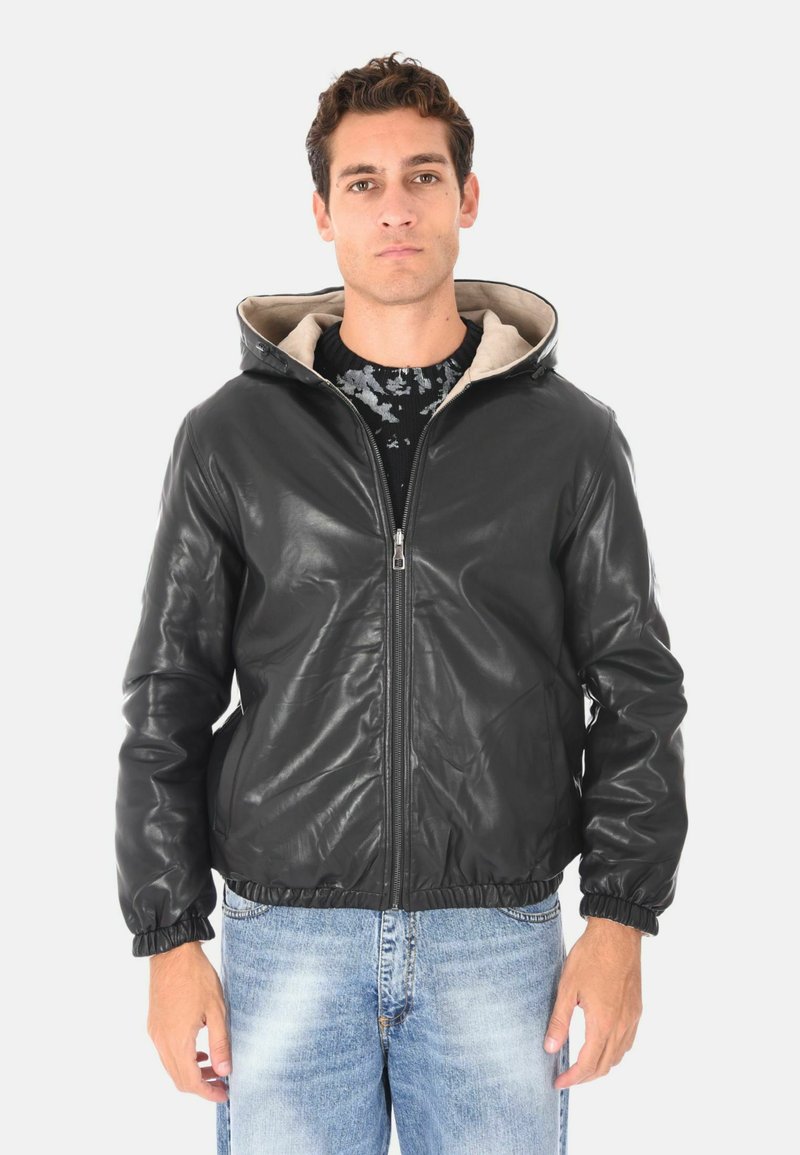 Uomo che indossa una giacca a zip nera in pelle con cappuccio sopra un maglione nero a fantasia e jeans chiari sbiaditi, in piedi di fronte a uno sfondo semplice.