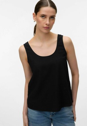 Jeune femme aux cheveux bruns attachés en queue de cheval, portant des boucles d'oreilles argentées en forme d'anneau, un haut noir sans manches et un jean bleu, devant un fond uni.