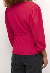 Rød, tekstureret bluse med puffede ærmer, tilpasset i taljen, parret med mørke jeans. Har en rund halsudskæring og subtile vandrette striber.