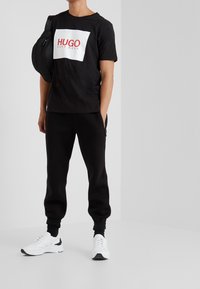 Sort bomulds T-shirt med et hvidt rektangulært logo, der viser "HUGO" i rødt. Sammen med sorte jogger-bukser og hvide sneakers.